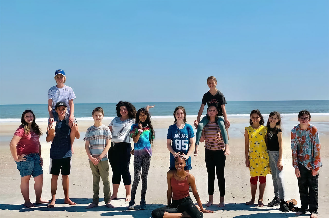 A group of teenage students standing and smiling together on the beach with the ocean behind them.