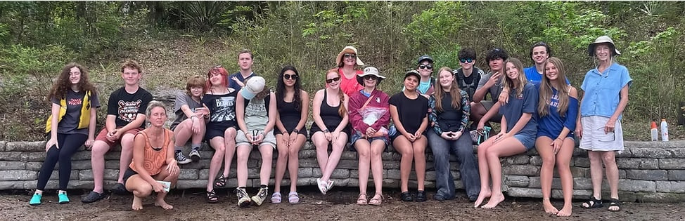 A large group of high-school students and a teacher gathered together, sitting and standing along a low stone wall in front of woodland.