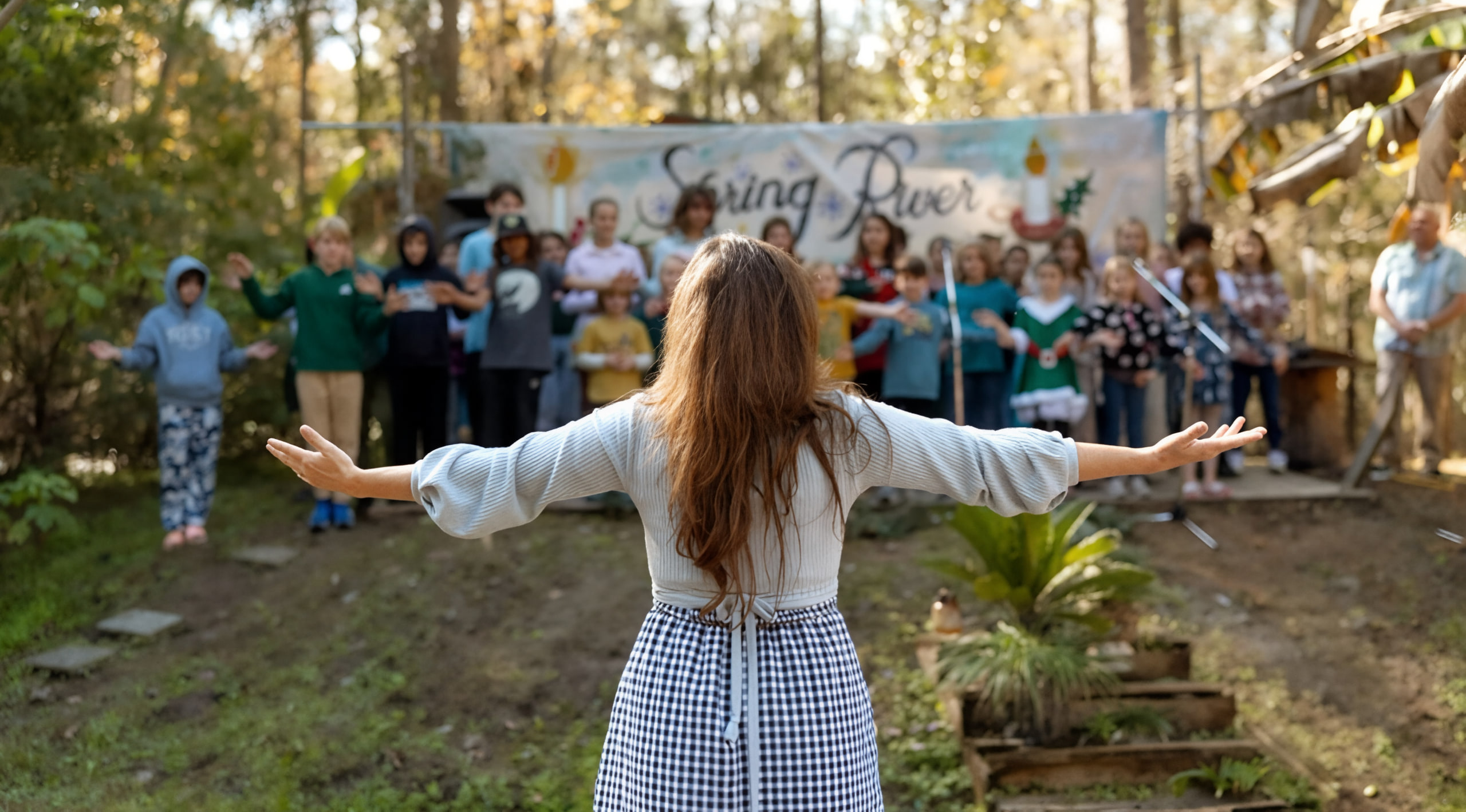 A teacher stands with arms open before a group of students gathered in an outdoor forest clearing beneath a hand-painted Spring River banner.