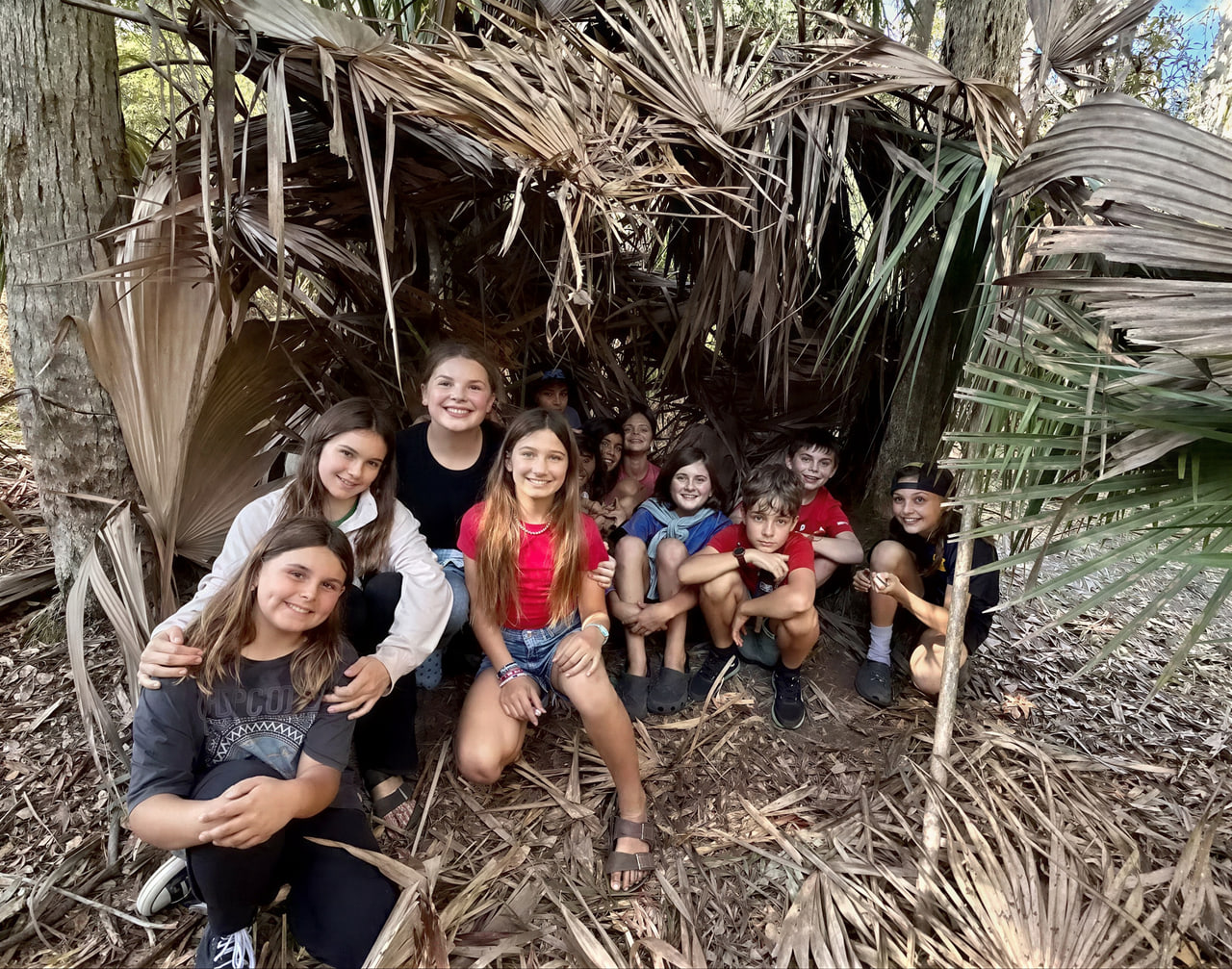 A group of children crouched together and smiling among tall green palmetto fronds in the woods.