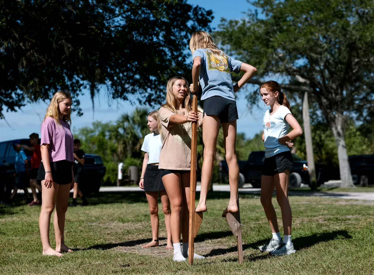 Middle-school students playing on wooden stilts and balance stakes on a sunny grassy field beneath live oak trees, one student balances on tall wooden stilts while classmates steady and watch her.